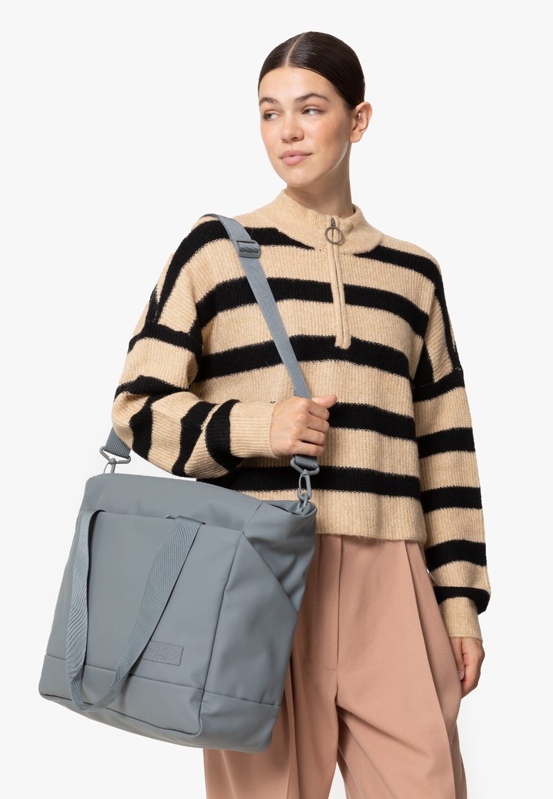 Sac fourre-tout gris avec une surface texturée, de forme rectangulaire, à doubles poignées et bandoulière ajustable. Porté avec un pull à rayures beige et noir.