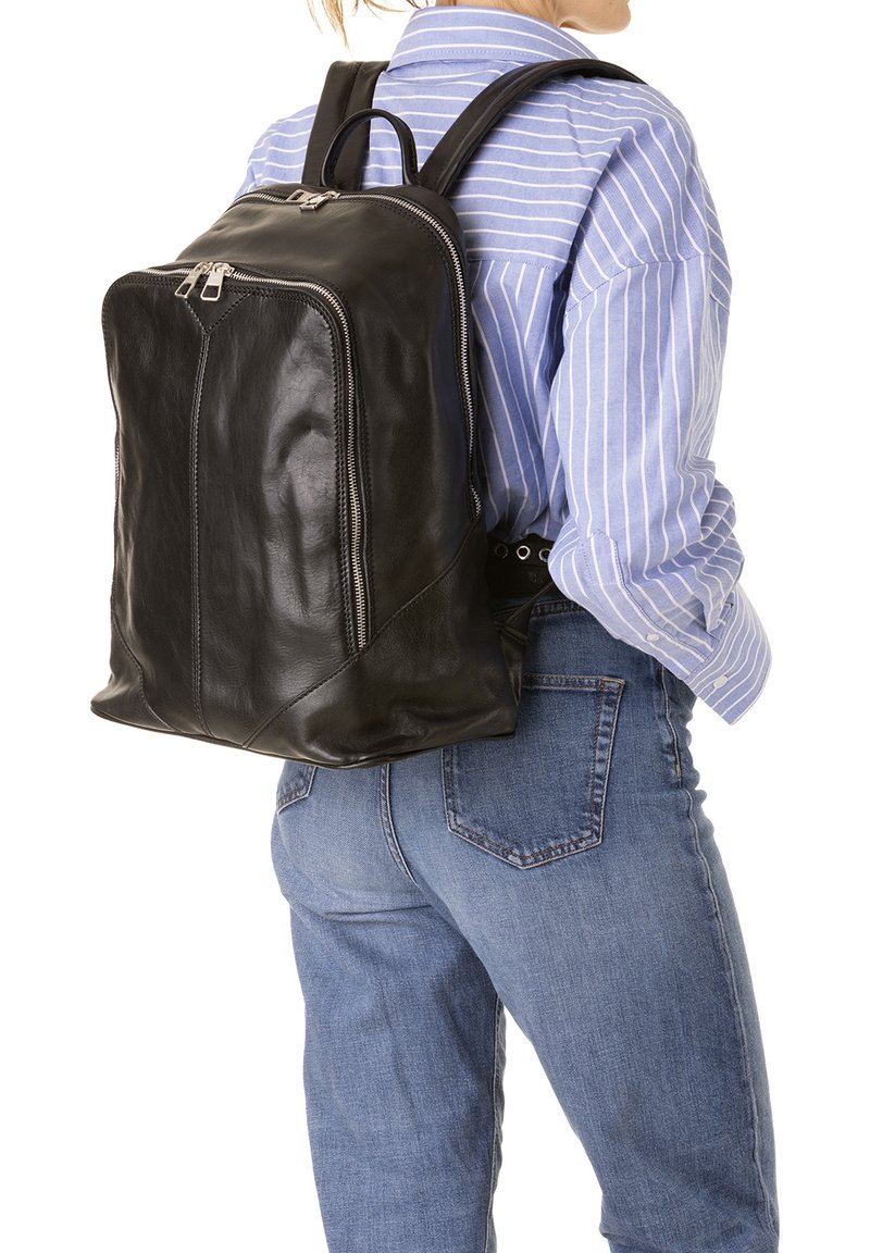 Un sac à dos en cuir noir avec une texture lisse, une fermeture à glissière sur le dessus et des bretelles réglables, porté sur une chemise à rayures bleues et un jean en denim.