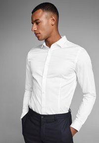 Witte blouse met lange mouwen en knopen, slim fit met een gladde textuur; gecombineerd met donkere broek; model in profiel tegen een grijze achtergrond.