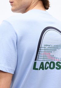 Νεαρός άνδρας που φοράει γαλάζιο μπλουζάκι με μεγάλο γραφικό ρακέτας τένις και μερικό κείμενο "LACOS" στην πλάτη.