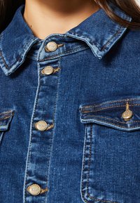 Jeansskjorta med spetsig krage, metallknappar i guldton och bröstfickor; mörkblått tyg med synliga sömmar och lätt slitage.