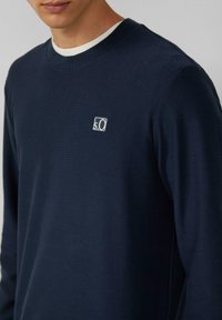 Pullover a maniche lunghe ribbed di colore blu navy con scollo rotondo e un piccolo logo bianco nella parte sinistra del petto. Tessuto morbido e texture piacevole.