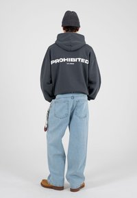 Grå hettegenser med „PROHIBERT“ i fete, hvite bokstaver på baksiden, kombinert med lyseblå jeans og brune sko. Myk tekstur, avslappet passform.