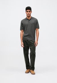 Een jonge man staat in een donkergrijze polo, donkergroene broek en tankleurige suède schoenen tegen een effen lichte achtergrond.