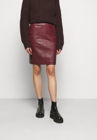 Bleistiftrock aus burgunderrotem Leder mit Seitennähten, kombiniert mit einem dicken, dunklen Strickpullover und schwarzen Stiefeletten mit dicker Sohle.