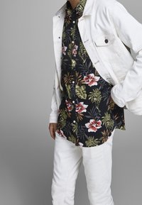 Vit denimjacka över en svart blommig skjorta med röda och gröna mönster, i kombination med vita byxor. Nära bild av överkroppen endast.