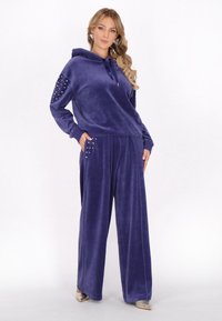 Hanorac din velur violet cu accente de mărgele pe mâneci și pantaloni cu croială largă asortați, prevăzuți cu buzunare. Textură fină și design ușor.