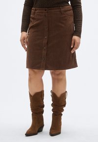 Fustă maro din catifea corespondență, cu închidere frontală cu nasturi și buzunare. Purtați cu cizme maro de cowboy până la gleznă, cu un toc ușor.