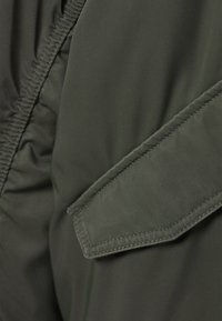 Gros plan sur un tissu vert foncé avec des coutures et une poche à rabat rectangulaire sur une veste ou un manteau.