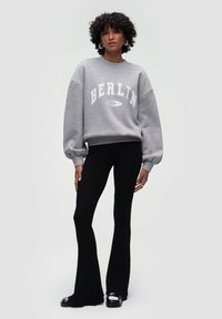 Szary sweter z napisem "BERLIN" w białym kolorze, w zestawieniu z czarnymi, żeberkowymi spodniami w stylu dzwonów. Materiały wydają się miękkie, z luźnym krojem i fakturowanymi detalami.