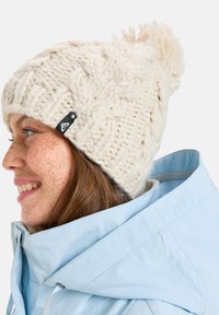 Gehaakte crèmekleurige beanie met een gestructureerd kabelpatroon en een pompon bovenop. Voorzien van een zwart etiket met een logo. Draagt een lichtblauwe jas.