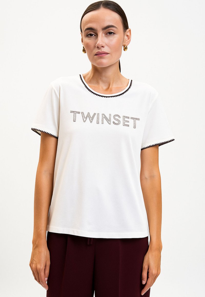 Donna che indossa una maglietta bianca a maniche corte con la scritta "TWINSET" e bordi neri su colletto e maniche, abbinata a pantaloni scuri.
