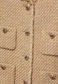 Beige gestructureerd jasje met goudkleurige knopen met kleurrijke accenten en franjes langs de randen en zakken.