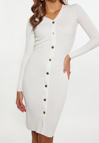 Vit ribbad klänning med V-ringning, långa ärmar, knappstängning framtill med svarta och gyllene knappar, samt en slits på sidan vid fållen.