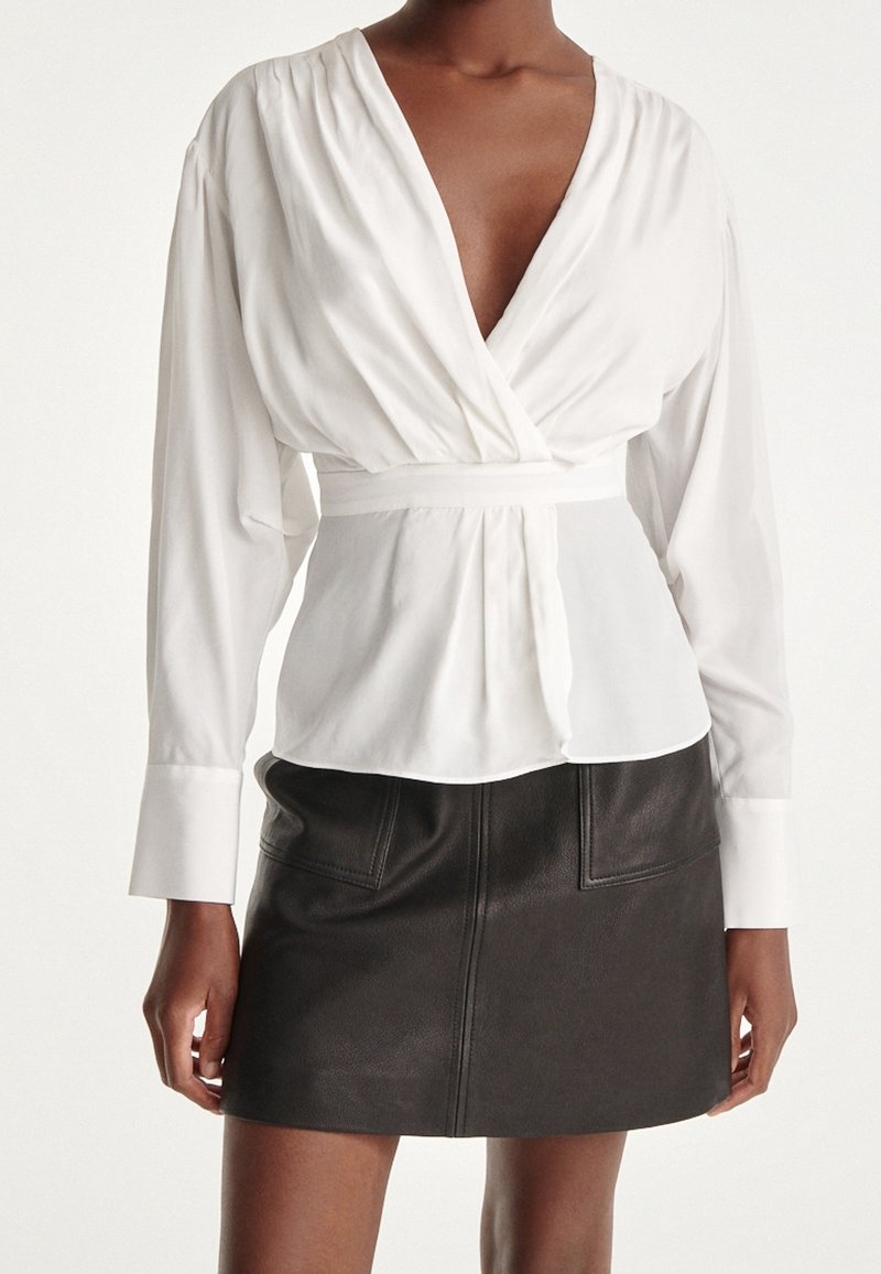 Vit omslagblus med djup V-ringning och långa ärmar, åtsittande vid midjan. Kombinerad med en svart läder minikjol med fickor.