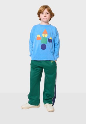 Helles blaues Langarmshirt mit buntem geometrischem Charakterdruck, kombiniert mit grüner Jogginghose mit Seitenstreifen und weißen Akzenten.