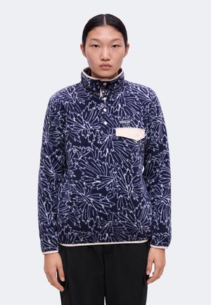 Giovane con capelli neri e lisci indossa un pile blu navy con motivo floreale, bordi beige e pantaloni neri.