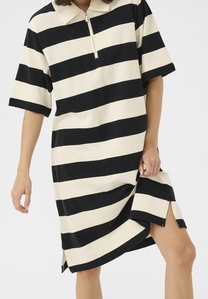 Personne portant une robe noire et blanche à larges rayures horizontales, manches courtes et col à fermeture éclair devant, soulevant légèrement l'ourlet.
