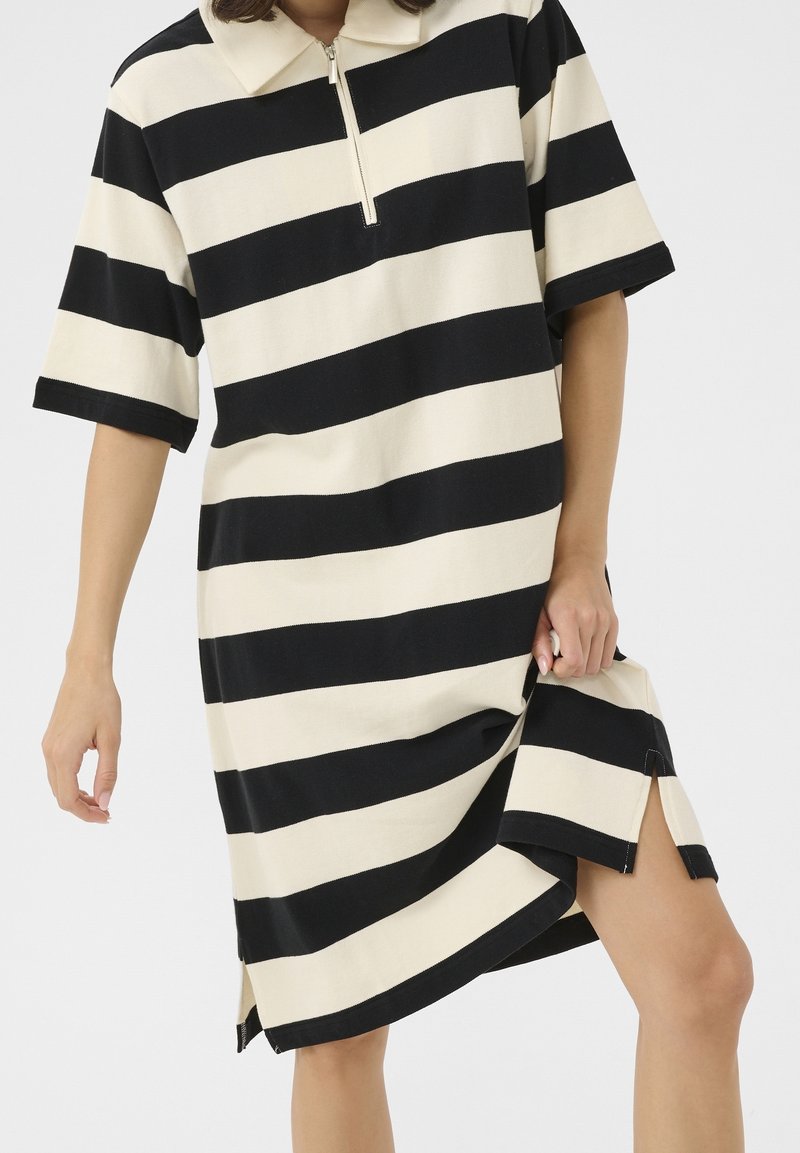 Personne portant une robe noire et blanche à larges rayures horizontales, manches courtes et col à fermeture éclair devant, soulevant légèrement l'ourlet.