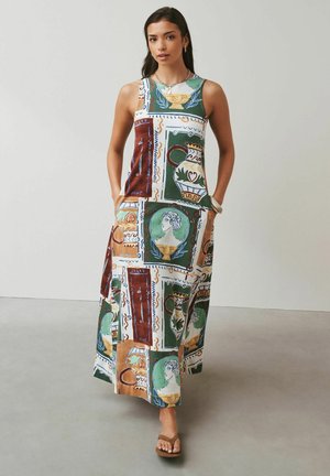 Femeie purtând o rochie fără mâneci, lungă până la glezne, cu imprimeu artistic colorat de vase și busturi clasice, stând în fața unui fundal simplu.