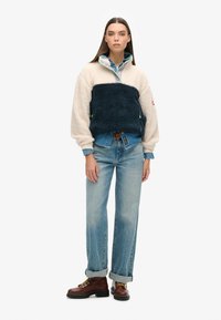 Pull en polaire avec une partie supérieure crème et une partie inférieure bleu foncé, doté d'un col zippé, d'accents verts, associé à un jean bleu ample et des bottes marron.