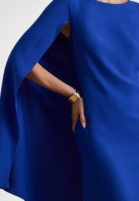 Királlykék ruha lebegő köpeny ujj dizájnnal, sima textúrával. Arany mandzsettagyűrű van a csuklón.