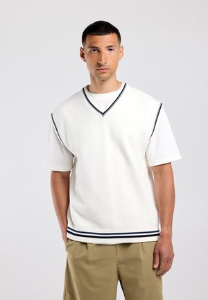 Jonge man met kort haar die een wit kort shirt draagt onder een witte gebreide vest met zwarte strepen en beige broek, staand tegen een effen achtergrond.