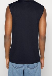 Man met een gebruinde huid die een mouwloos zwart shirt en lichtblauwe jeans draagt, gezien van achteren tegen een effen witte achtergrond.