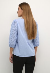 Haut bleu clair avec un dos uni et des manches bouffantes texturées présentant un motif en zigzag et des découpes en œillets, associé à un pantalon noir.