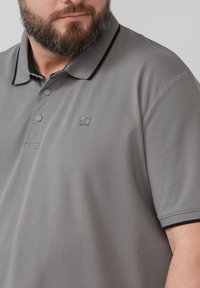 Szara koszulka polo z ciemnoniebieskimi detalami przy kołnierzyku, wykonana z teksturowanej tkaniny. Posiada małe logo na piersi oraz podwójny zamek na guziki.