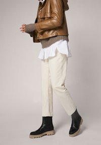 Veste en cuir marron courte, pantalon beige à jambes larges, bottines noires à semelles épaisses. Une chemise blanche superposée dépasse du pull.