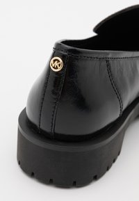 Chaussure en cuir noir avec une finition texturée, une semelle plateforme épaisse noire et un accent logo MK doré près du talon.