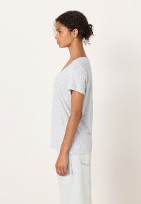 Jauna moteris su garbanotais plaukais surištais į kuodą, dėvinti šviesiai pilk ą trumpomis rankovėmis V formos kaklo palaidinę ir šviesius džinsus, stovinti šonu.