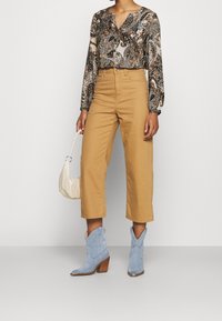 Musterbluse mit langen Ärmeln in Braun- und Blautönen, kombiniert mit hochtaillierten, tanfarbenen, weit geschnittenen Hosen. Hellblaue Wildleder-Stiefeletten.