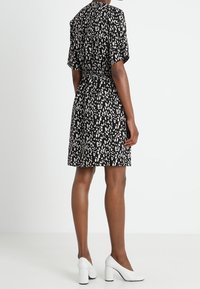 Vestido negro con manchas abstractas blancas, mangas cortas, cintura ajustada y longitud por encima de la rodilla, combinado con zapatos de tacón cuadrado blancos.