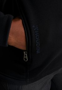 Musta vetoketjullinen takki pehmeällä tekstuurilla. Takissa on voimakas "Houdini"-logo tummansinisellä brodeerauksella vetoketjun lähellä. Lähikuva taskusta.