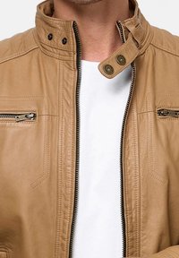 Hellbraune Lederjacke mit hohem Kragen, Reißverschlusstaschen und Druckknopfverschluss. Glatte Textur und minimalistische Designdetails.