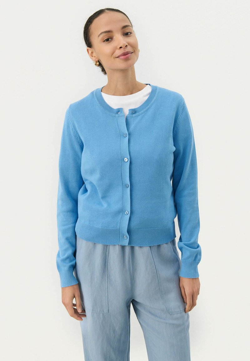 Cardigan azzurro chiaro realizzato in materiale morbido, con scollo rotondo, chiusura a bottoni, polsini e orlo a costine. Indossato sopra una camicia bianca.