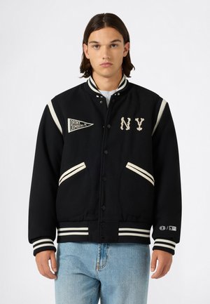Giovane uomo che indossa una giacca varsity nera con strisce bianche, toppe "NY" e "Bronx Bombers", e jeans azzurri, in piedi davanti a uno sfondo semplice.