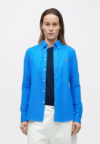 Svetlomodrá košeľa s dlhými rukávmi z pletenej tkaniny, s golierom na gombíky, prednými gombíkmi a malým logom. Nosená cez tmavomodrú košeľu.