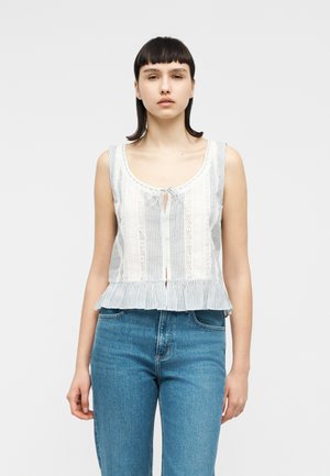 Noor naine, kes kannab valget ja sinist triibulist varrukateta pluusi pitsdetailidega ning siniseid kõrge vöökohaga teksapükse, seisab ühevärvilise tausta ees.