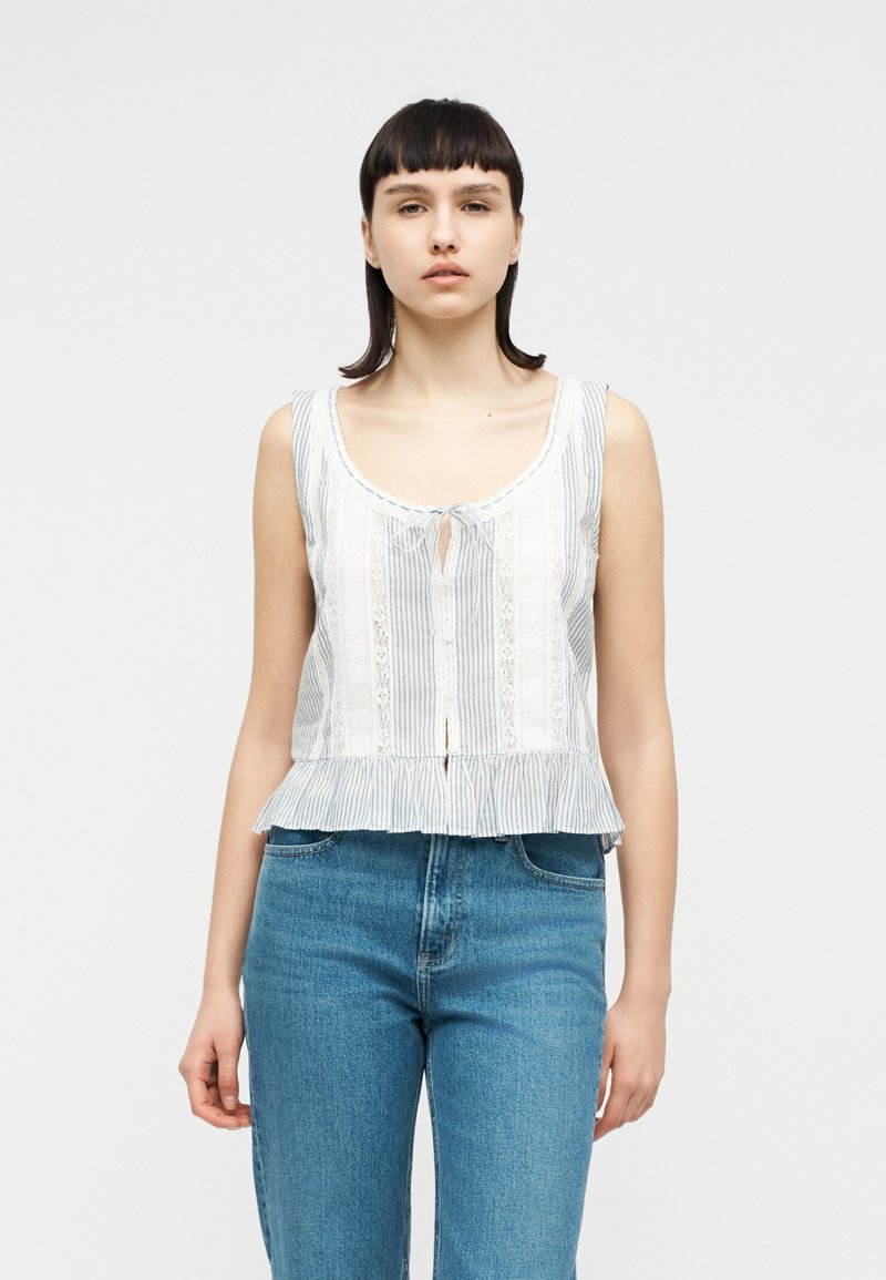 Νεαρή γυναίκα που φοράει ένα αμάνικο λευκό και μπλε ριγέ μπλουζάκι με δαντελένιες λεπτομέρειες και μπλε τζιν με ψηλή μέση, στέκεται μπροστά σε ένα απλό φόντο.