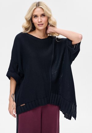 Blondynka w luźnym, czarnym dzianinowym ponczo z podwiniętymi rękawami i bordowymi spodniami, pozująca z jedną ręką za szyją.