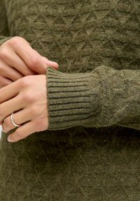 Hands adjusting the cuff of an olive green knitted sweater with a textured pattern, one hand wearing a simple silver ring on the ring finger.