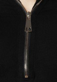 Czarna bluza z kapturem na zamek błyskawiczny w kolorze złotym. Posiada prążkowaną teksturę wzdłuż kołnierza oraz zbliżenie na szczegóły zamka.