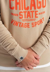Felpa di un leggero beige con testo arancione audace che riporta "CHICAGO," "STATE" e "VINTAGE SPORT." Maniche a coste e una piccola etichetta con il logo visibile.