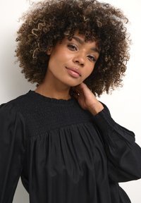 Schwarze Bluse mit langen Ärmel und gerafftem Oberteil, runder Ausschnitt und gerafftem Stoff. Lockiges Haar sorgt für Volumen vor einem neutralen Hintergrund.