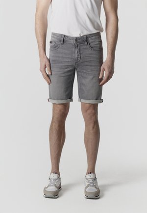 Mand iført op rullede grå denimshorts, hvide sneakers og en hvid t-shirt, stående foran en ensfarvet baggrund.