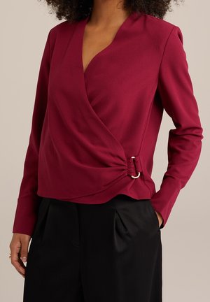 Mujer vistiendo un top de manga larga estilo cruzado color burdeos con detalle de hebilla de anillo plateado, combinado con pantalones negros plisados.