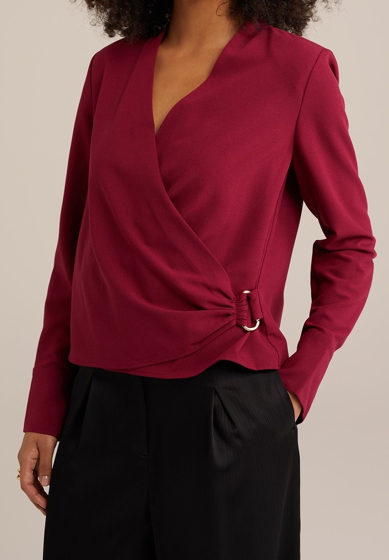 Femme portant un haut manches longues style cache-cœur bordeaux avec un détail de boucle anneau argenté, associé à un pantalon plissé noir.
